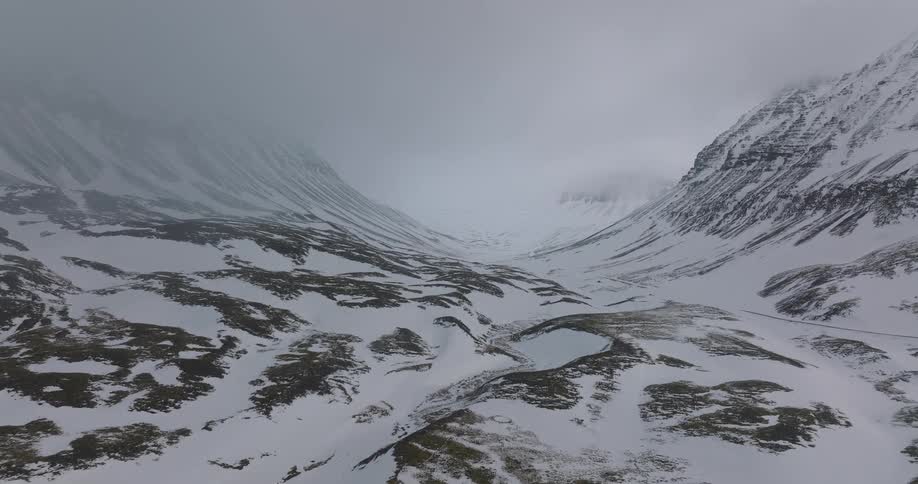 An aerial view slowly drifts forward through a vast, snow-covered mountain valley on a cold and foggy winter day