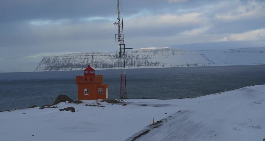 An aerial view slowly approaches a vibrant orange lighthouse perched on a snowy cliff overlooking a cold Icelandic fjord