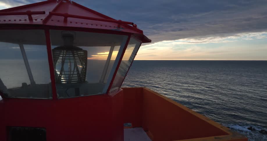 A closeup of a red lighthouse's Fresnel lens slowly pulls back to reveal a dramatic ocean sunset