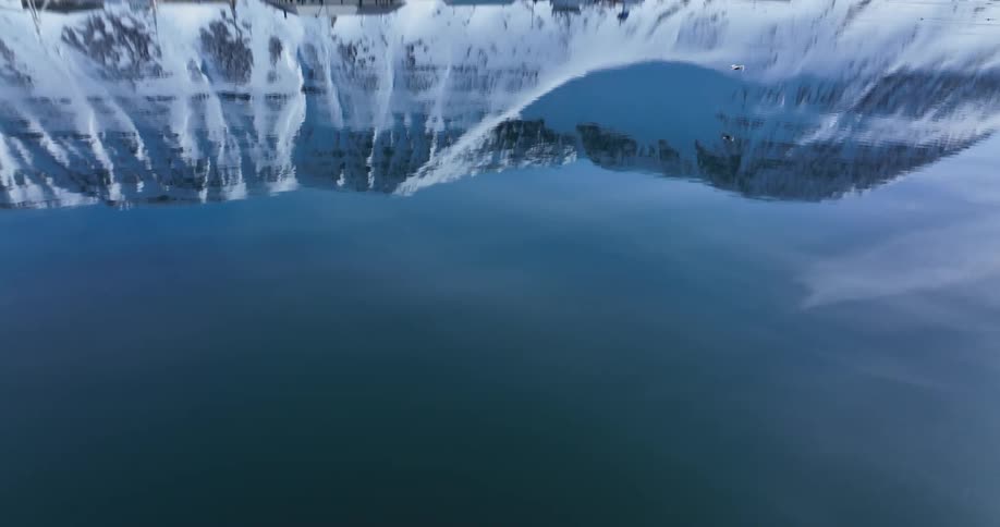 An inverted aerial shot reveals the surreal, mirror-like reflection of snowy arctic mountains on calm fjord waters as birds fly past