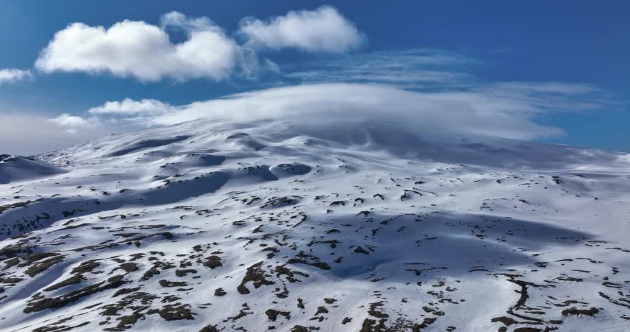 A majestic snow-covered mountain range sits under a deep blue sky as clouds flow over the peaks in a serene, looping aerial view