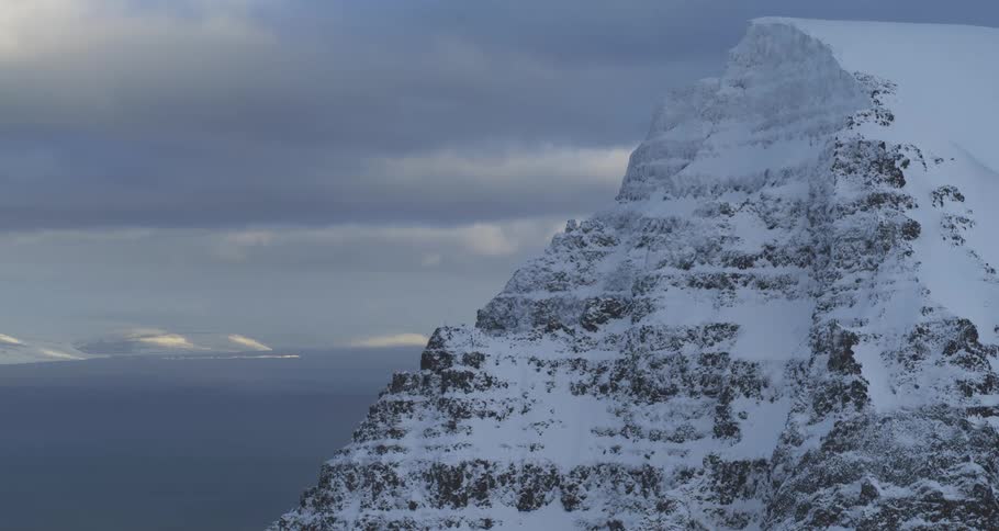 Majestic snow-covered mountain overlooks a vast ocean under a dramatic, cloudy sky in Iceland