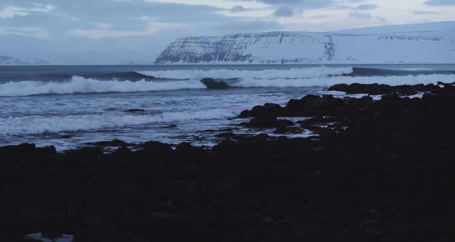 Slow motion waves crash on a dark, rocky coast with a snow-covered mountain in the background