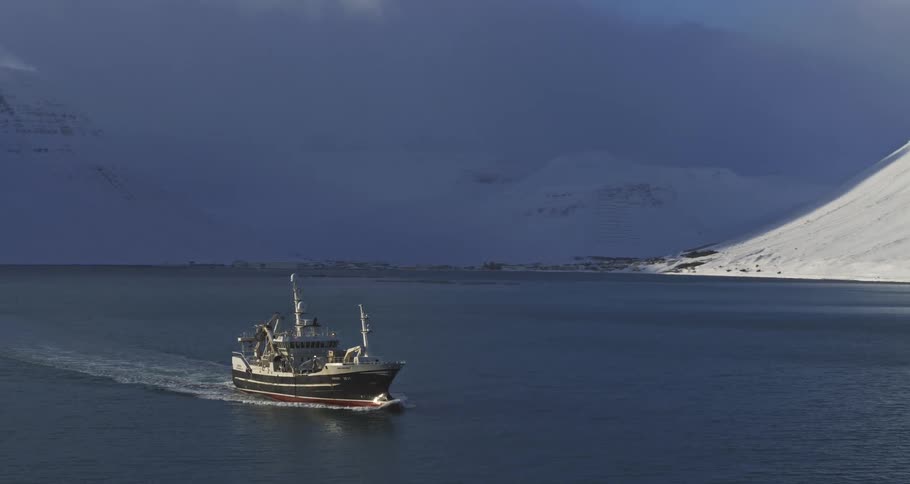 A fishing boat sails through an arctic fjord surrounded by snow-covered mountains as dramatic light breaks through the clouds