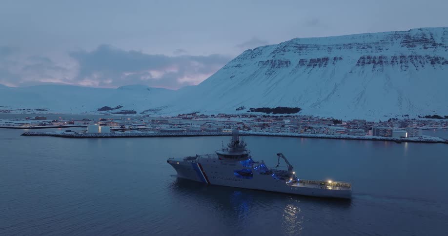 An Icelandic Coast Guard ship sits peacefully in a harbor at dusk with a snowy mountain landscape and a coastal town in the background