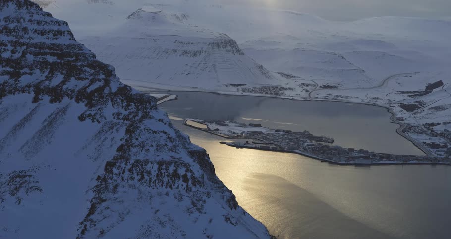 A majestic wide shot of a snowy fjord in Iceland, where a small coastal village sits on a peninsula as the sun reflects on the calm water