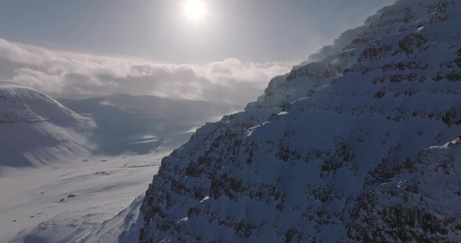 A slow pull-back aerial shot reveals a vast, sunlit, snow-covered mountain range as strong winds blow across the frigid landscape