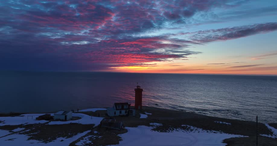 Aerial view of a striking red lighthouse on a snowy coast during a vibrant, colorful sunset over the ocean