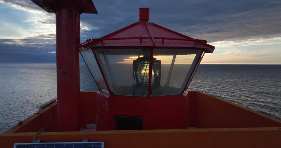 A slow pull-back shot reveals the intricate Fresnel lens inside a red lighthouse lantern room overlooking the ocean at sunset