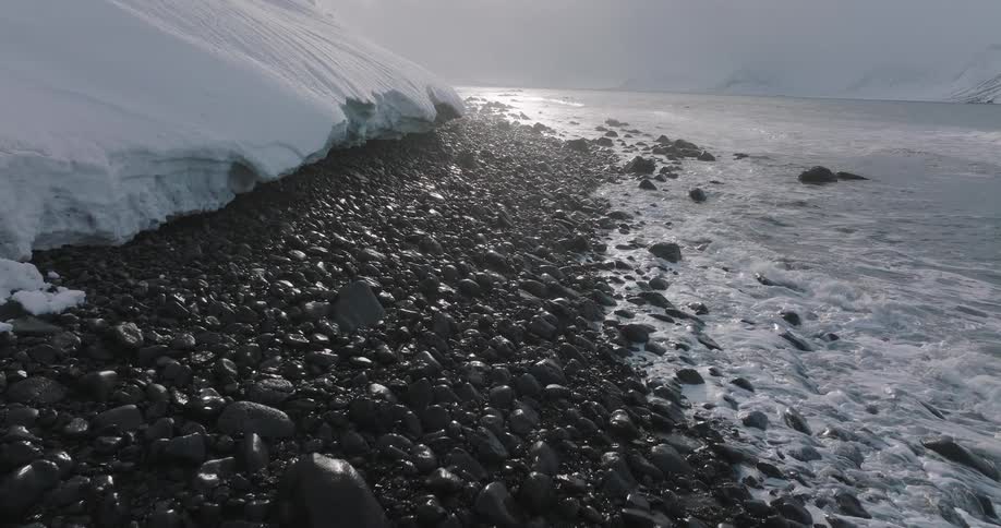 A slow forward shot over a rocky, arctic shoreline where gentle waves meet the snow-covered land