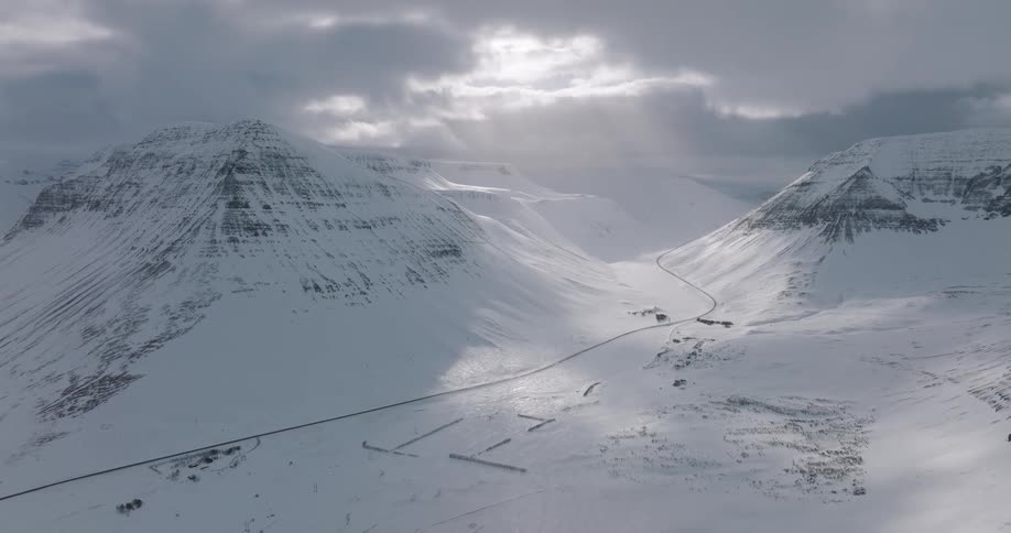 An epic aerial pullback reveals a vast, snowy mountain valley where sunbeams pierce through dramatic clouds
