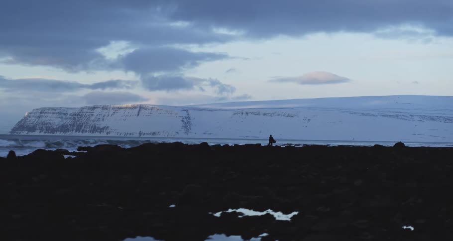 A lone surfer walks along a dark, rocky shoreline, preparing to enter the frigid ocean against a stunning backdrop of snow-covered mountains in Iceland