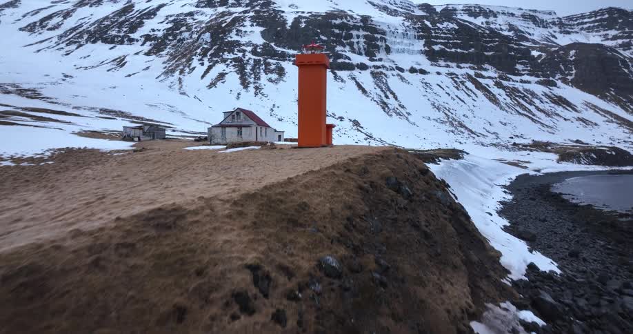 An aerial drone shot flies towards a bright orange lighthouse on a remote Icelandic coastline with snow-covered mountains in the background