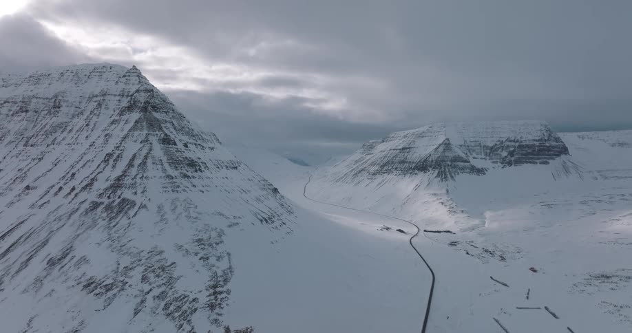 A dramatic aerial view of a remote road winding through a vast, snow-covered mountain valley under an overcast sky