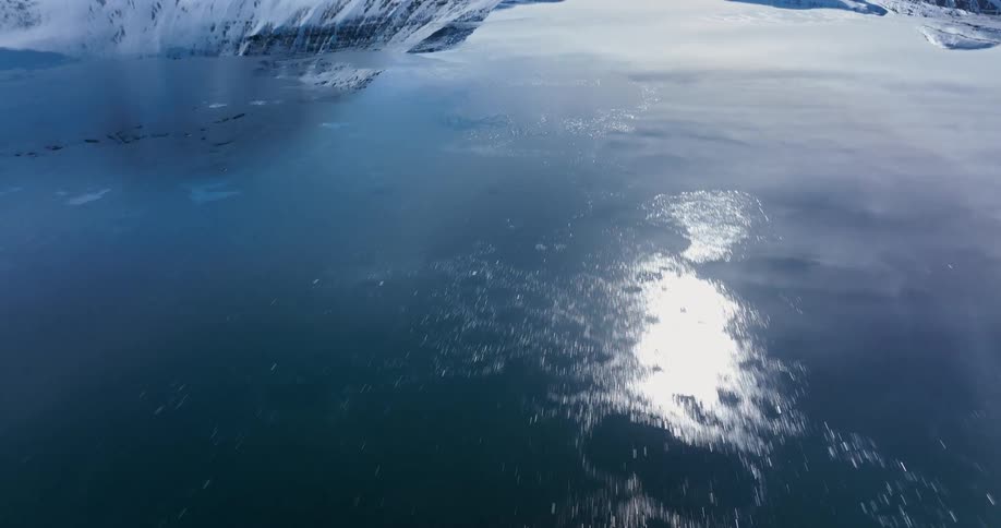 A surreal inverted time-lapse shows the sun and snowy mountains reflected on the surface of a rapidly freezing and thawing arctic fjord