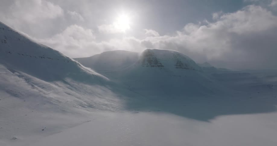 A static shot captures a vast, snow-covered mountain range as wind blows snow across the landscape under a hazy sun