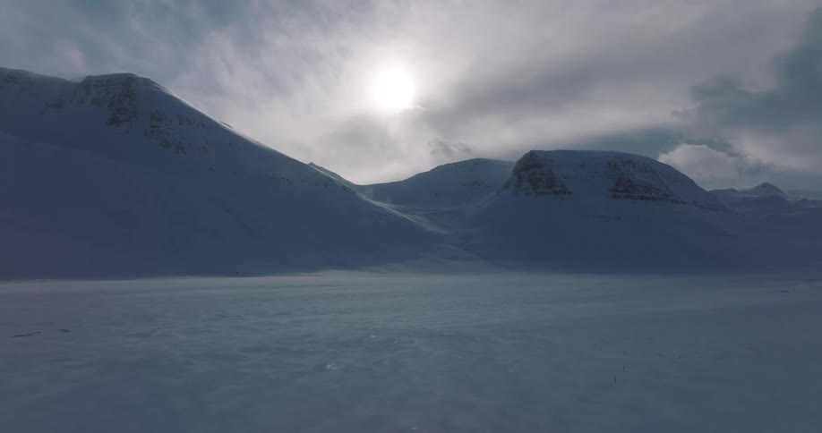 A stationary shot of blowing snow across a frozen valley surrounded by mountains under a hazy sun