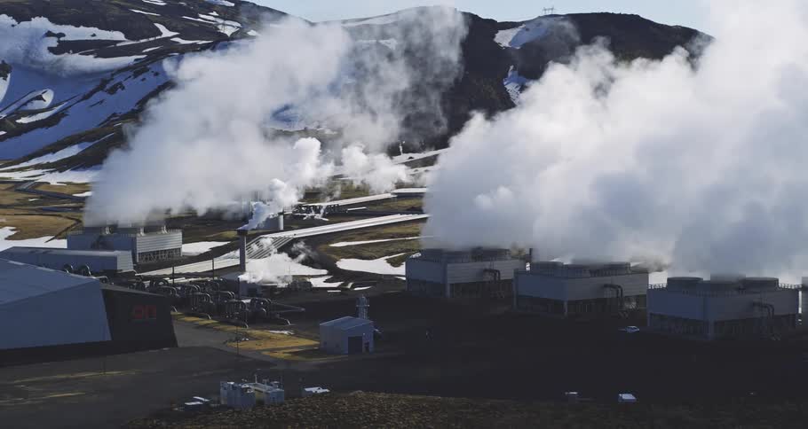 A wide aerial shot shows a geothermal power plant releasing plumes of steam with a backdrop of snowy mountains