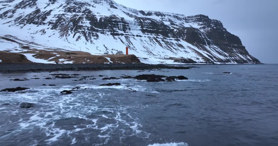 An aerial view flies over a cold ocean towards a remote lighthouse at the base of a snow-covered mountain on a cloudy day
