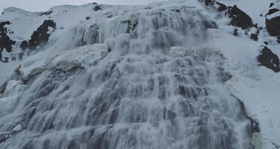 A powerful waterfall thunders down a steep, snow-covered mountainside in a dramatic, icy landscape