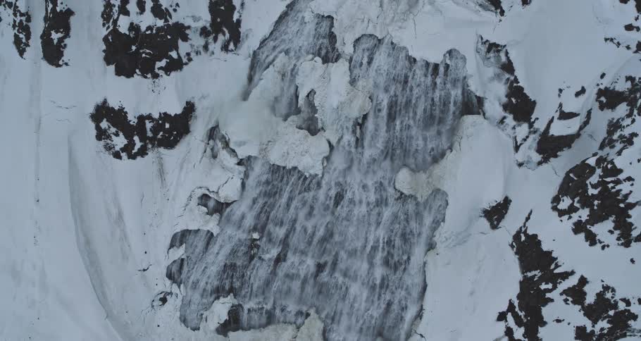 Ascending aerial top-down view of a powerful icy waterfall cascading through a snowy mountain landscape