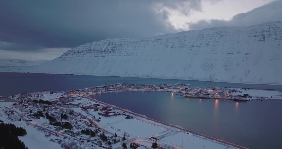 A stunning aerial view captures a snowy coastal town in Iceland, nestled beside a majestic mountain at twilight