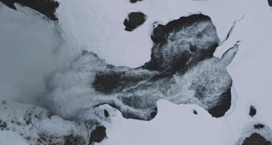 Top-down aerial shot reveals a raging river and waterfall cutting through a snow and ice-covered landscape