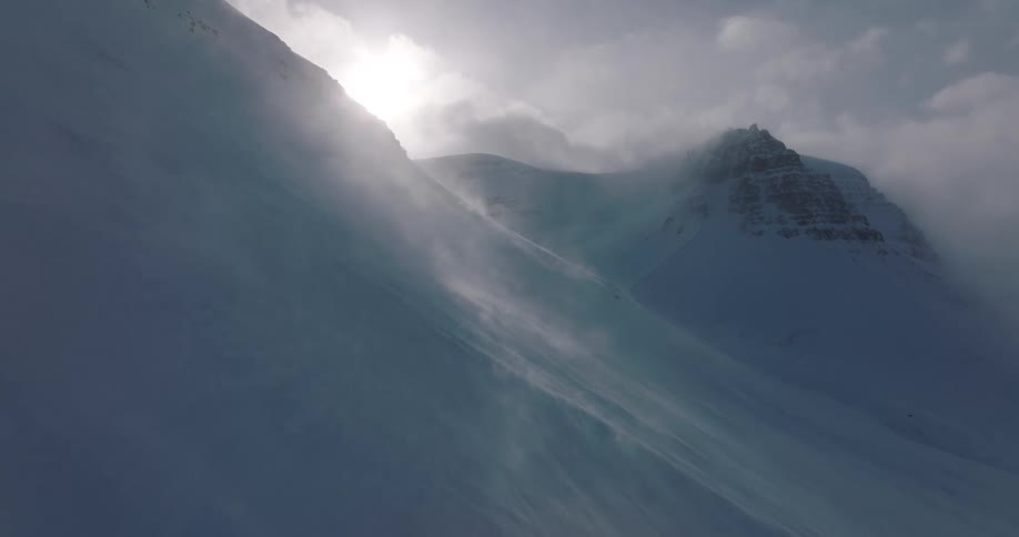 Blizzard winds blow snow across a stark, desolate mountain landscape with the sun breaking through the clouds