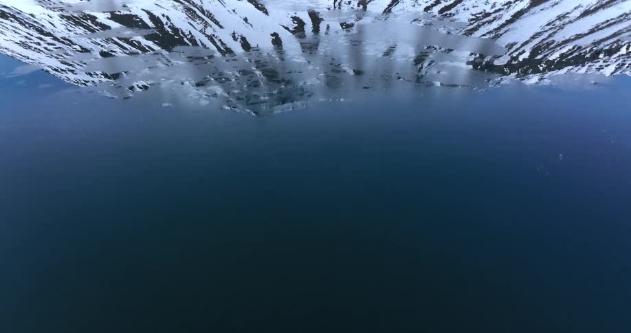 An inverted aerial view slowly flies over a calm lake, revealing the pristine reflection of snow-covered mountains