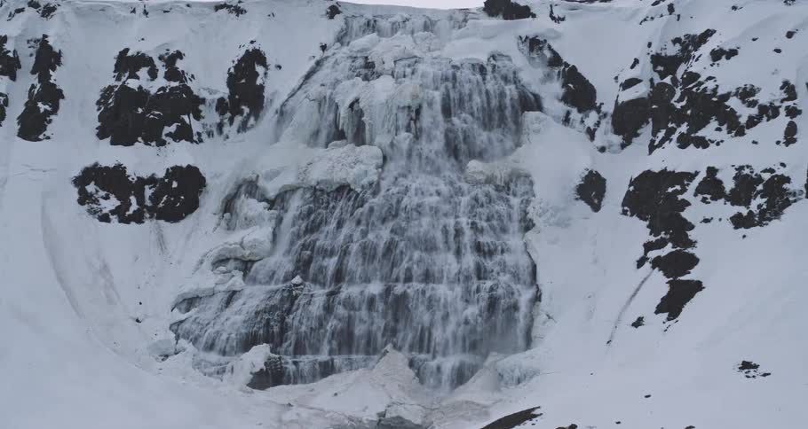 A stunning aerial view zooms in on a majestic, partially frozen waterfall cascading down a snow-covered mountain in a vast, wintry landscape