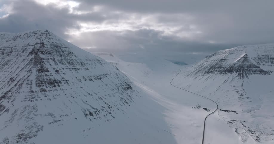 A lone highway winds through a majestic, snow-covered mountain valley under a dramatic, cloudy sky