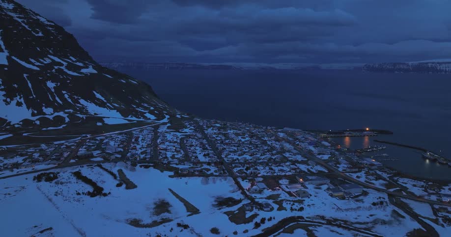 A slow aerial push over a serene snow-covered fishing village nestled between a mountain and the sea at night