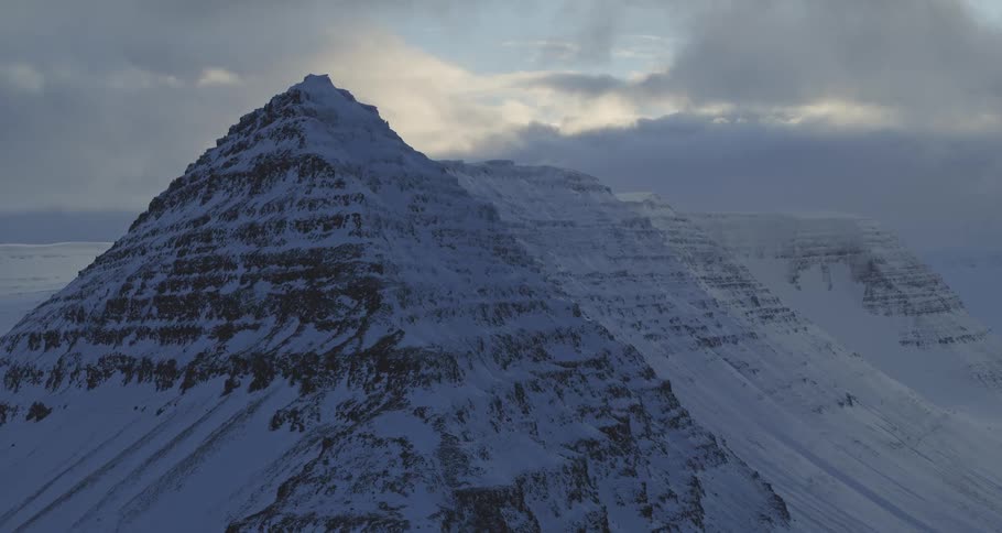 A majestic, snow-covered mountain looms large under a cloudy sky as the camera slowly zooms in on its peak