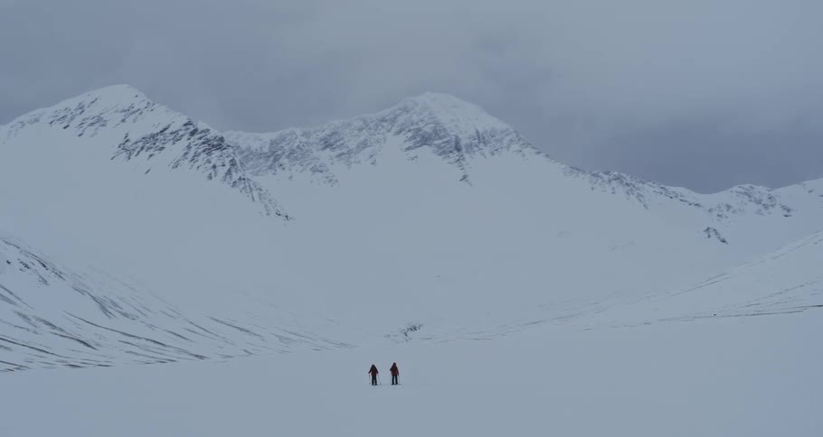 A slow zoom reveals two explorers dwarfed by the immense, snow-covered mountains of a vast, frozen valley