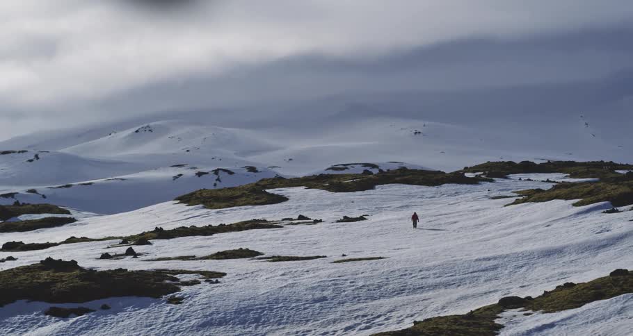 A solitary hiker in a red jacket treks across a vast and desolate snow-covered mountain landscape