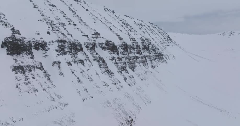 An epic aerial shot moves slowly along the windswept face of a vast, snow-covered mountain in Iceland