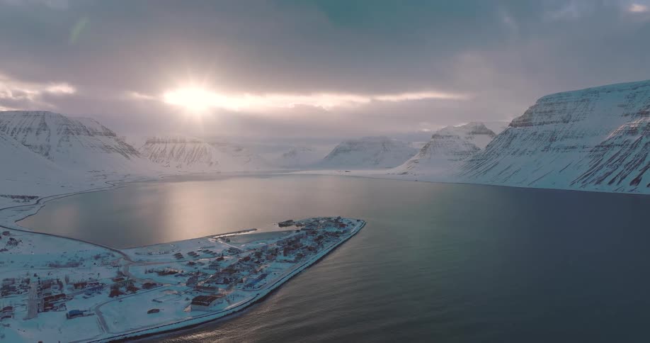 A serene aerial view shows a sunbeam breaking through clouds over a remote, snow-covered fishing village in a majestic fjord