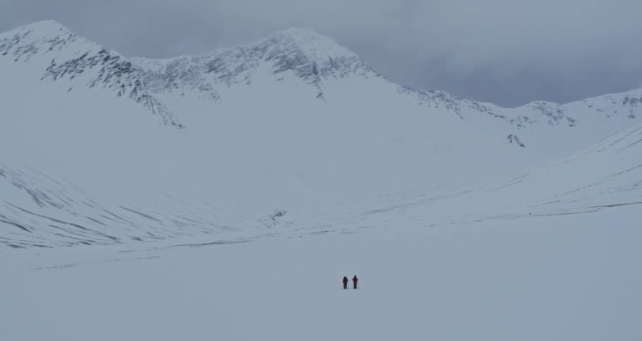 A panoramic shot reveals two tiny figures of explorers standing in the middle of a vast, white, snow-covered valley surrounded by imposing mountains