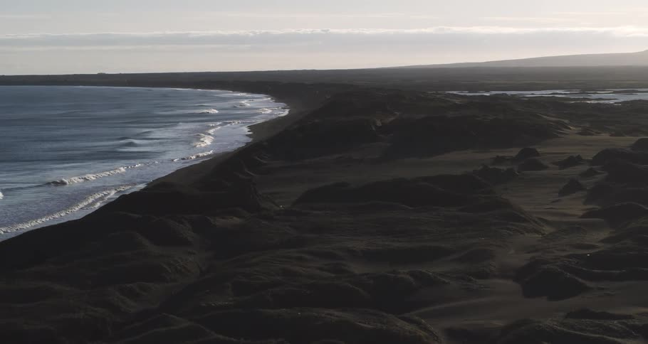An expansive high-angle view of a desolate black sand beach with dunes as ocean waves roll onto the shore under a hazy sky