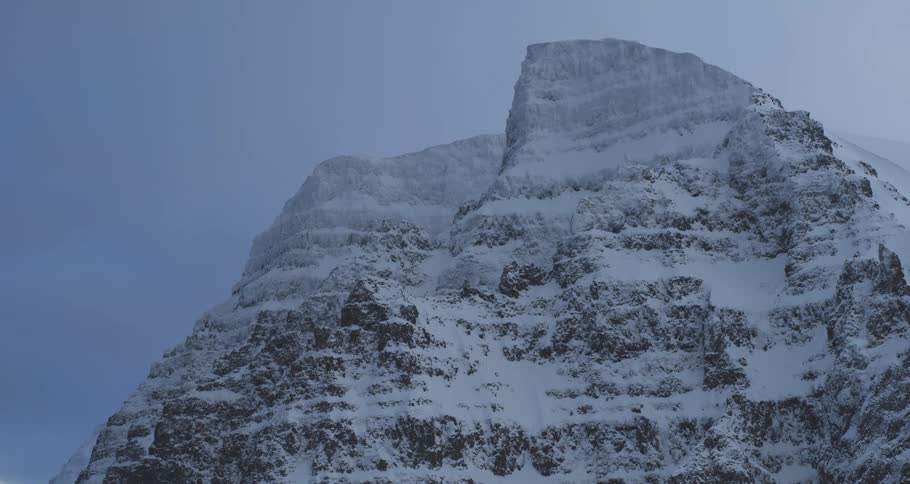 A low-angle static shot of snow blowing across a rugged, snow-covered mountain peak on a cold, overcast day