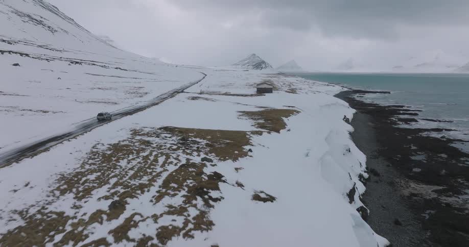 An aerial view follows a lone car traveling down a remote coastal road through a vast, snow-covered landscape with mountains and the sea in the distance