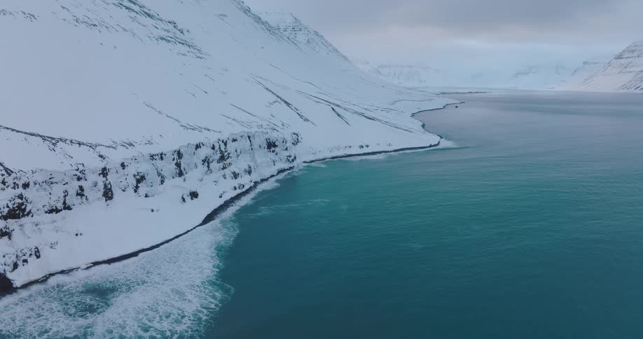 Calm turquoise waves wash ashore on a remote, snow-covered coastline nestled between vast mountains in a winter landscape