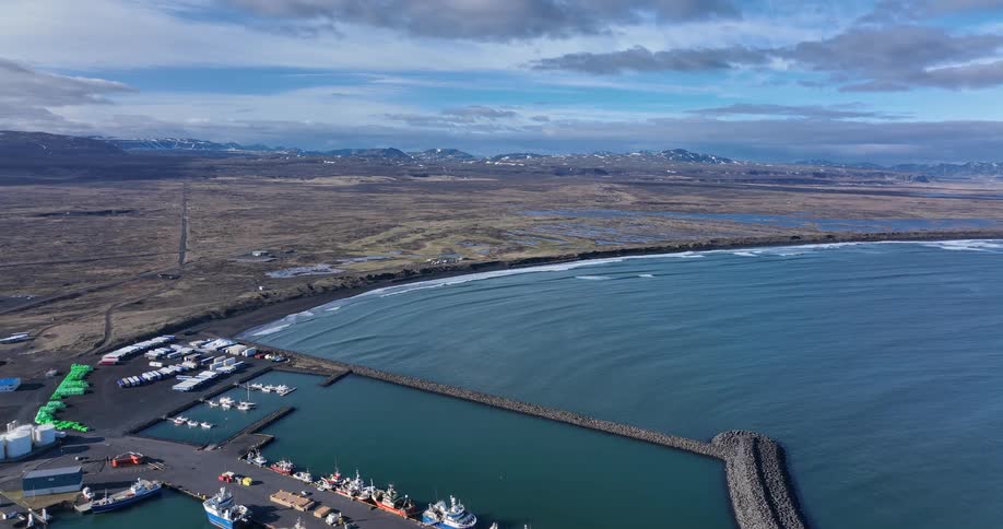 Sweeping aerial view of a fishing harbor on a dramatic Icelandic coastline with distant snow-capped mountains