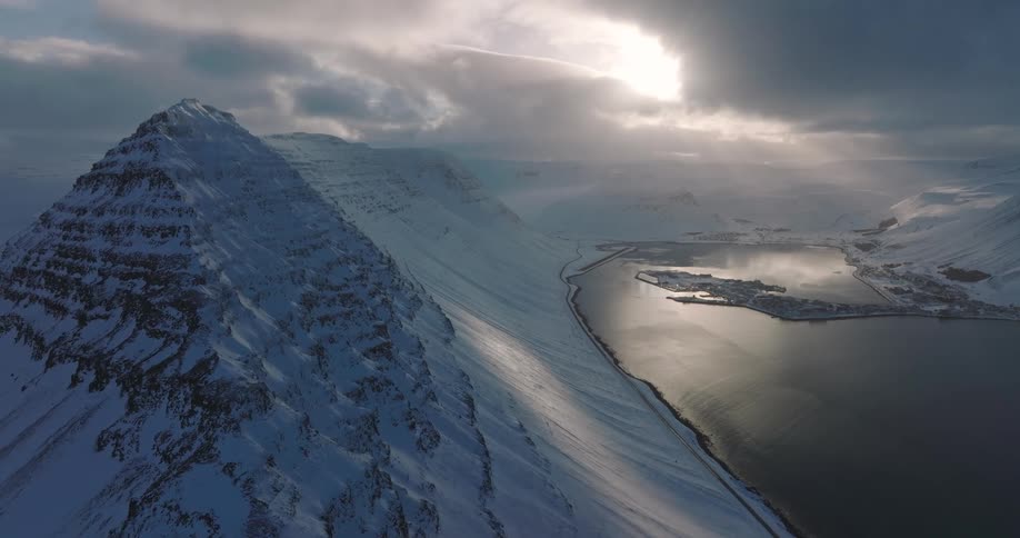 An epic aerial view of a snow-covered mountain overlooking a coastal village in a vast Icelandic fjord under a dramatic, cloudy sky