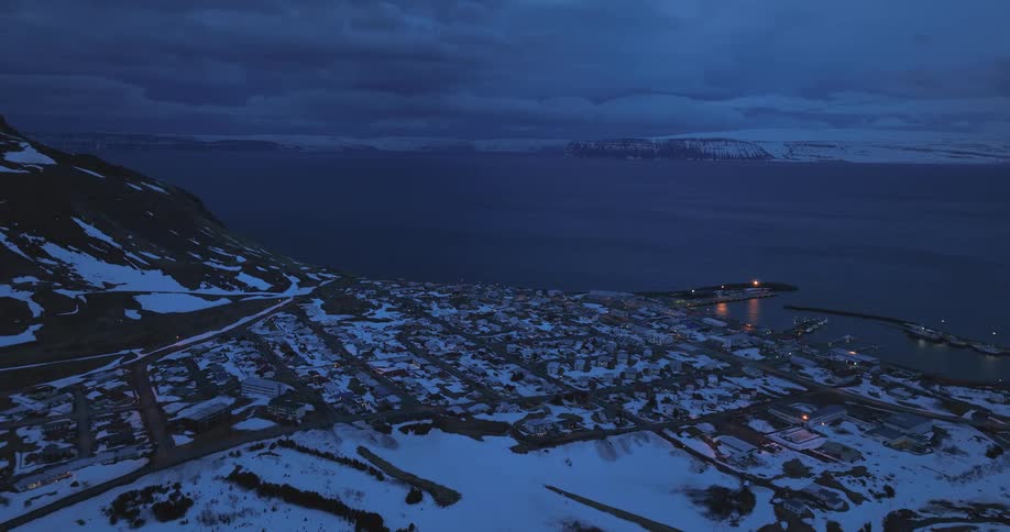 A slow aerial view reveals a quiet, snow-covered coastal village glowing with lights during the blue hour in Iceland