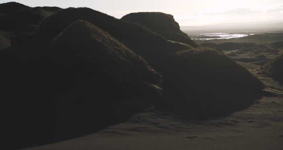 A slow, cinematic drone shot glides over windswept black sand dunes under a hazy, setting sun