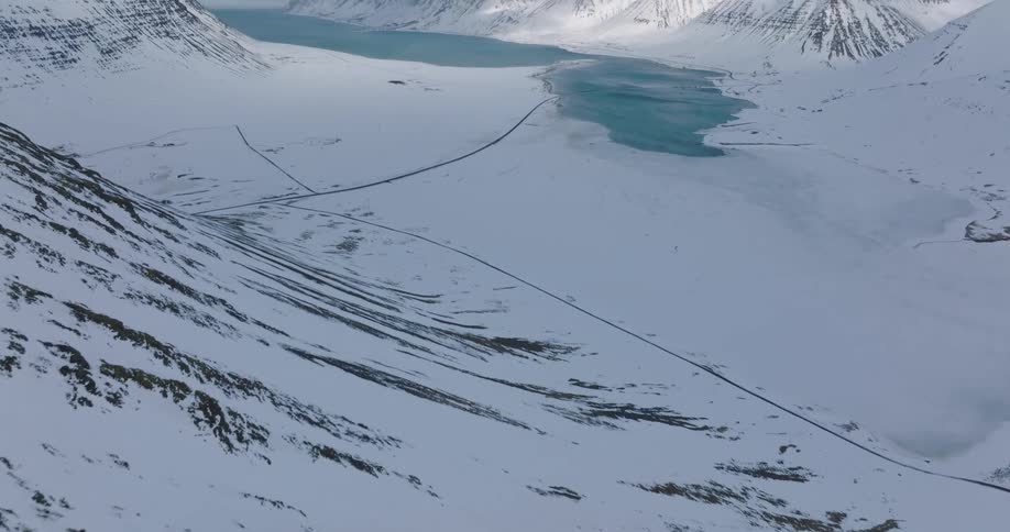 Slow aerial pullback revealing a vast, snow-covered Icelandic fjord surrounded by majestic mountains under a cloudy sky