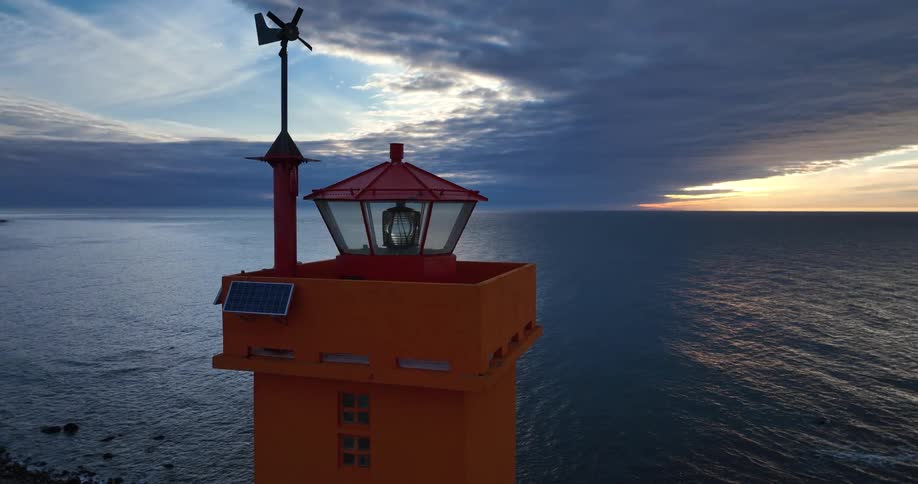 An aerial view captures a striking orange lighthouse with its light rotating against a dramatic sunset sky over the ocean