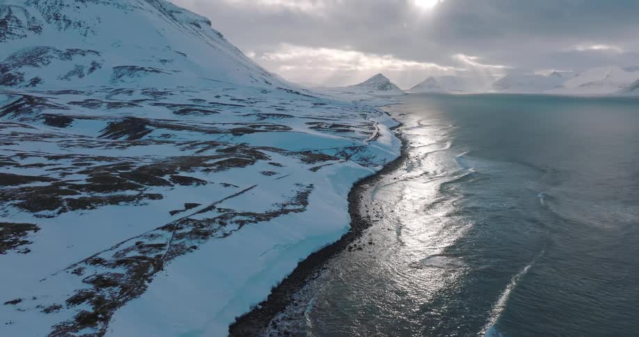 A stunning aerial view pans up to reveal a dramatic, snow-covered coastline with waves crashing ashore as sunbeams break through the clouds over distant mountains