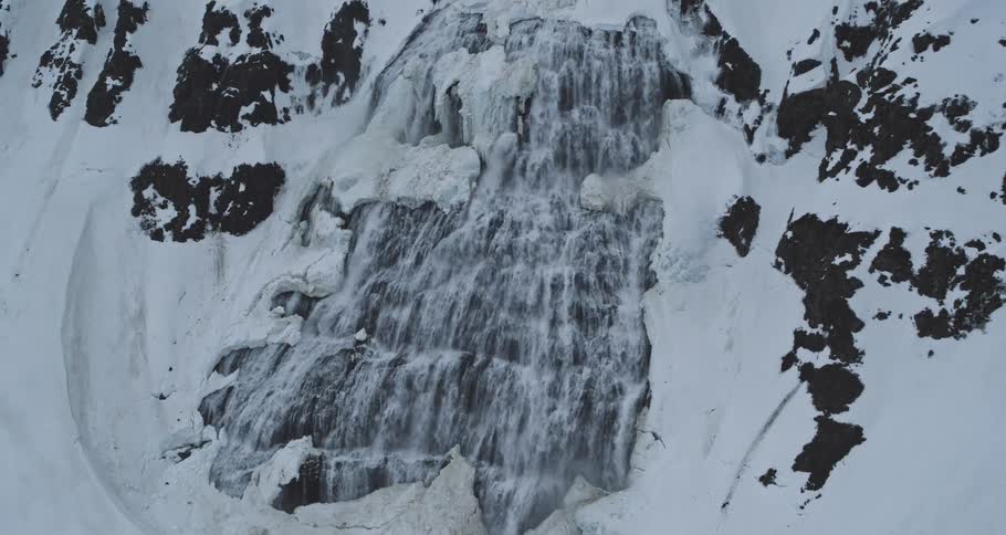 A drone slowly pulls back to reveal a majestic waterfall cascading down a vast, snow-covered mountainside in winter